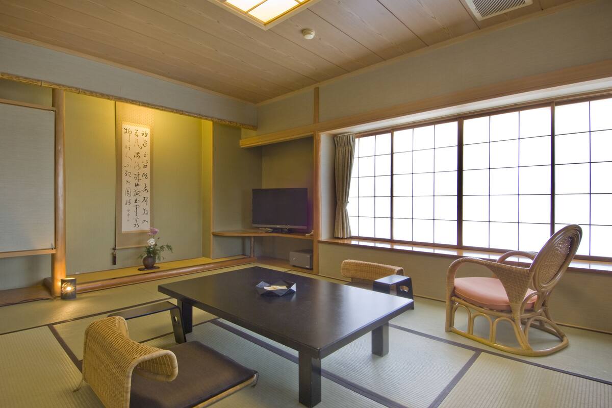 A traditional Japanese room with tatami flooring, a low table, a television, and a window with a view.