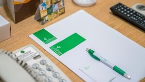 A desk with a white notepad, a green pen, a calculator, a black remote control, and a small stand with brochures. - LeGreen Suite Senayan (Jakarta)