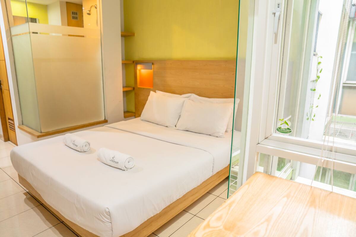 A hotel room with a bed, a wooden headboard, a glass shower enclosure, a bedside table, and a window with a view of greenery.