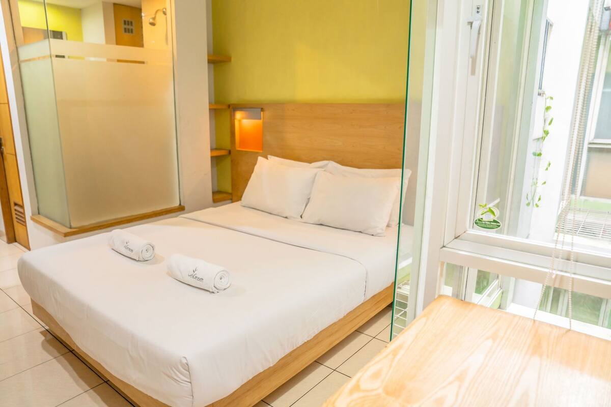 A hotel room with a bed, a wooden headboard, a glass partition, a nightstand with a lamp, and a window with a view.