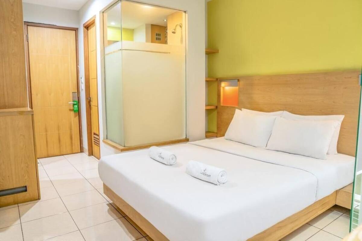 A hotel room with a bed, a wooden wardrobe, a mirror, and a wall-mounted shelf with a lamp.