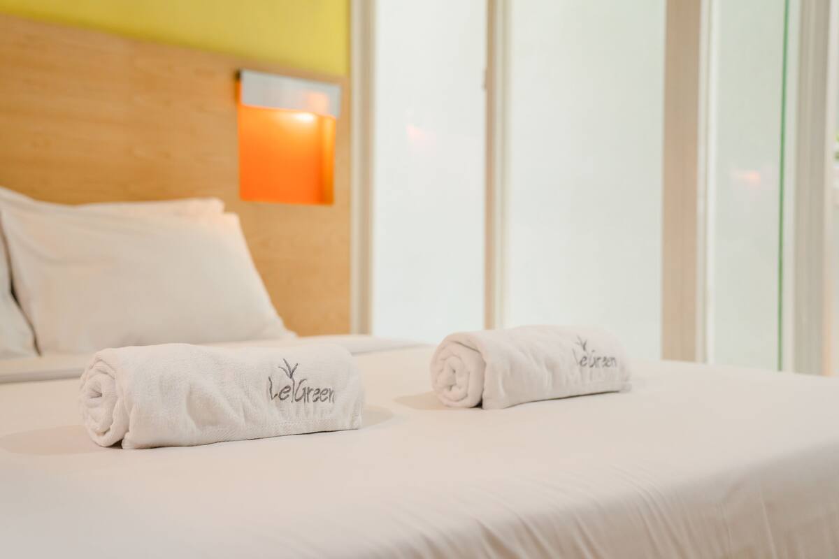 A hotel room with a neatly made bed, two rolled towels with the hotel name, a wooden headboard, and a wall-mounted lamp.