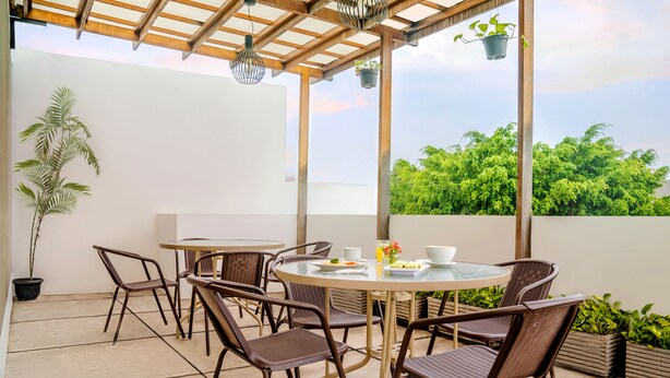 Une terrasse extérieure couverte, avec une table ronde et des chaises, des plantes en pot et une vue sur la verdure.