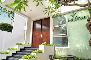 Welcome to LeGreen, where lush greenery surrounds the entrance, featuring a wooden door and stairs leading to a covered seating area.