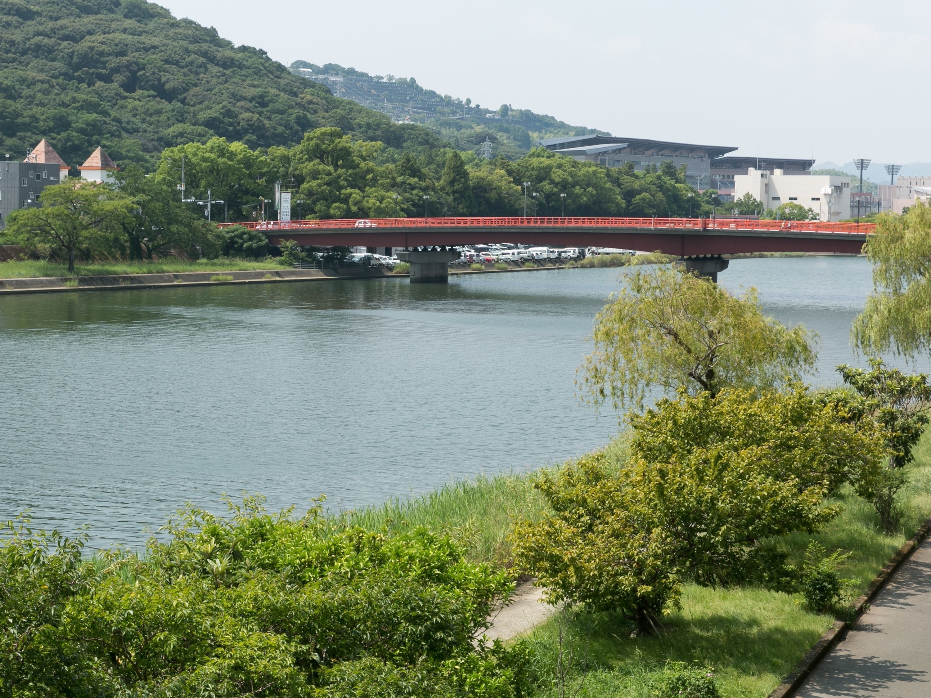 A river with a red bridge, lush greenery, and a path alongside the river.