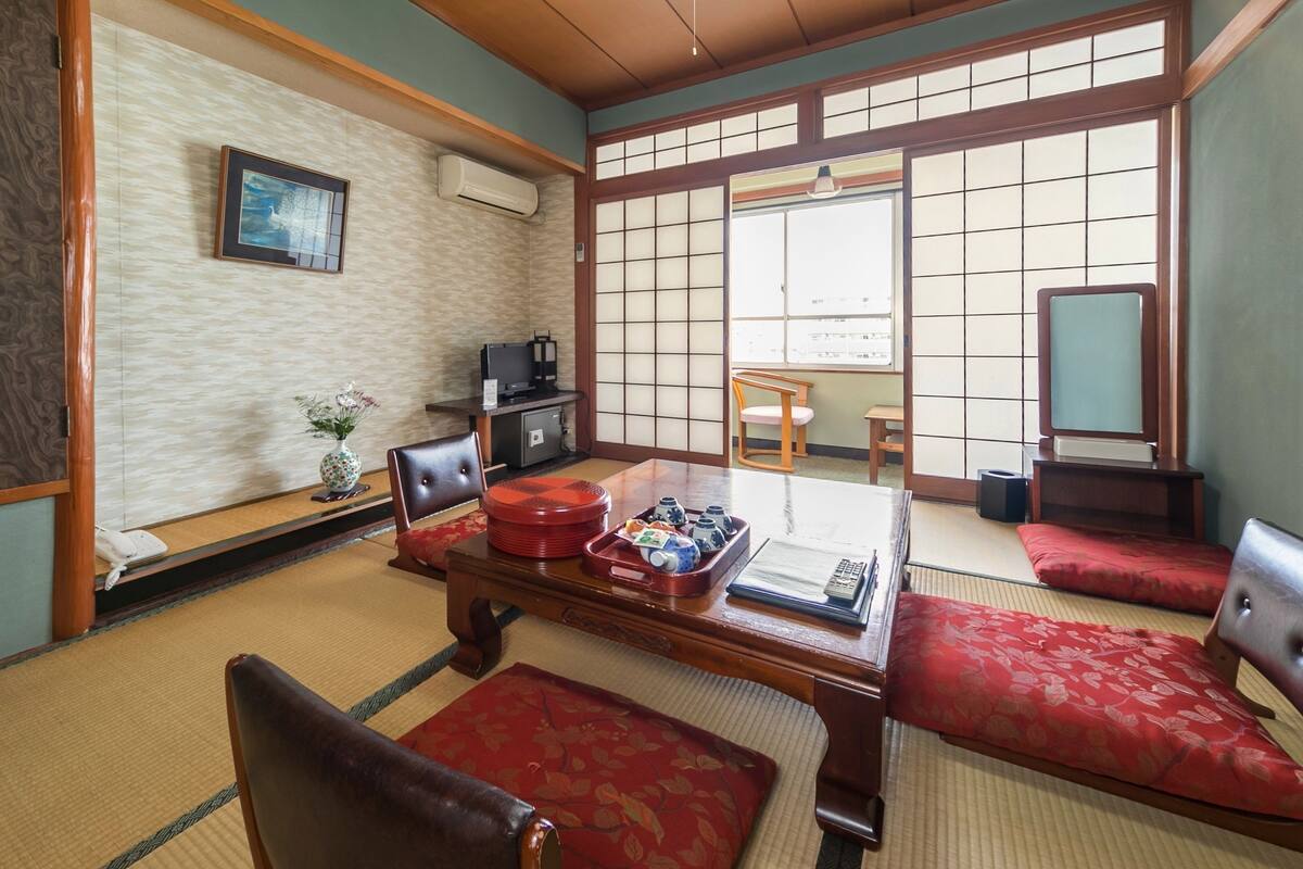 Um quarto japonês tradicional com tatames, uma mesa de madeira baixa, almofadas vermelhas, uma televisão e uma porta deslizante com vista para o exterior.