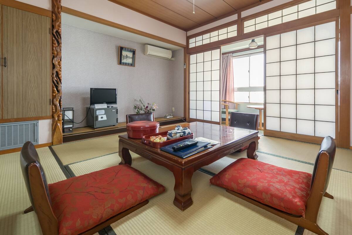 Um quarto japonês tradicional com tatames, uma mesa de madeira baixa com almofadas, uma televisão e um painel deslizante shoji.