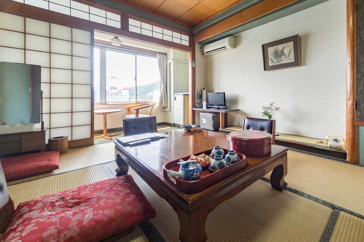 Um quarto japonês tradicional com tatames, uma mesa baixa com jogo de chá, um assento almofadado vermelho, uma televisão e vista para prédios através de um shoji.