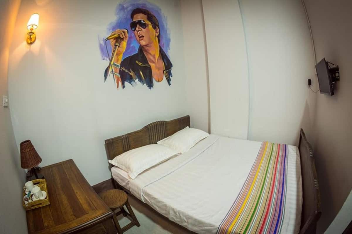 A hotel room with a bed, a wooden nightstand, a lamp, a wall-mounted TV, and a painting.