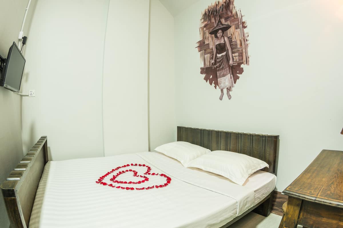 A neatly made bed with a heart-shaped decoration of rose petals, a wooden nightstand with a lamp, and a wall-mounted artwork.