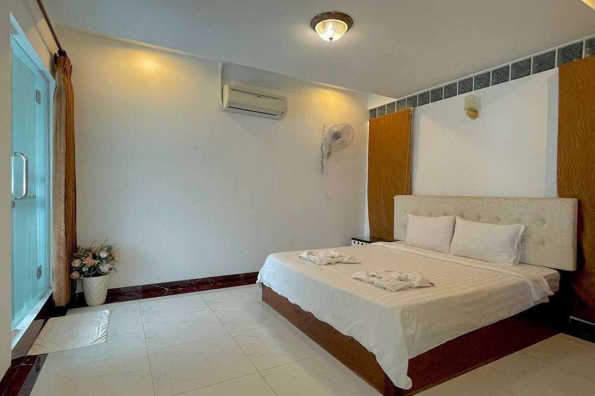 Ein Hotelzimmer mit einem Bett, einer Klimaanlage, einem Wandventilator, einer Zimmerpflanze und einer Tür mit Vorhang.