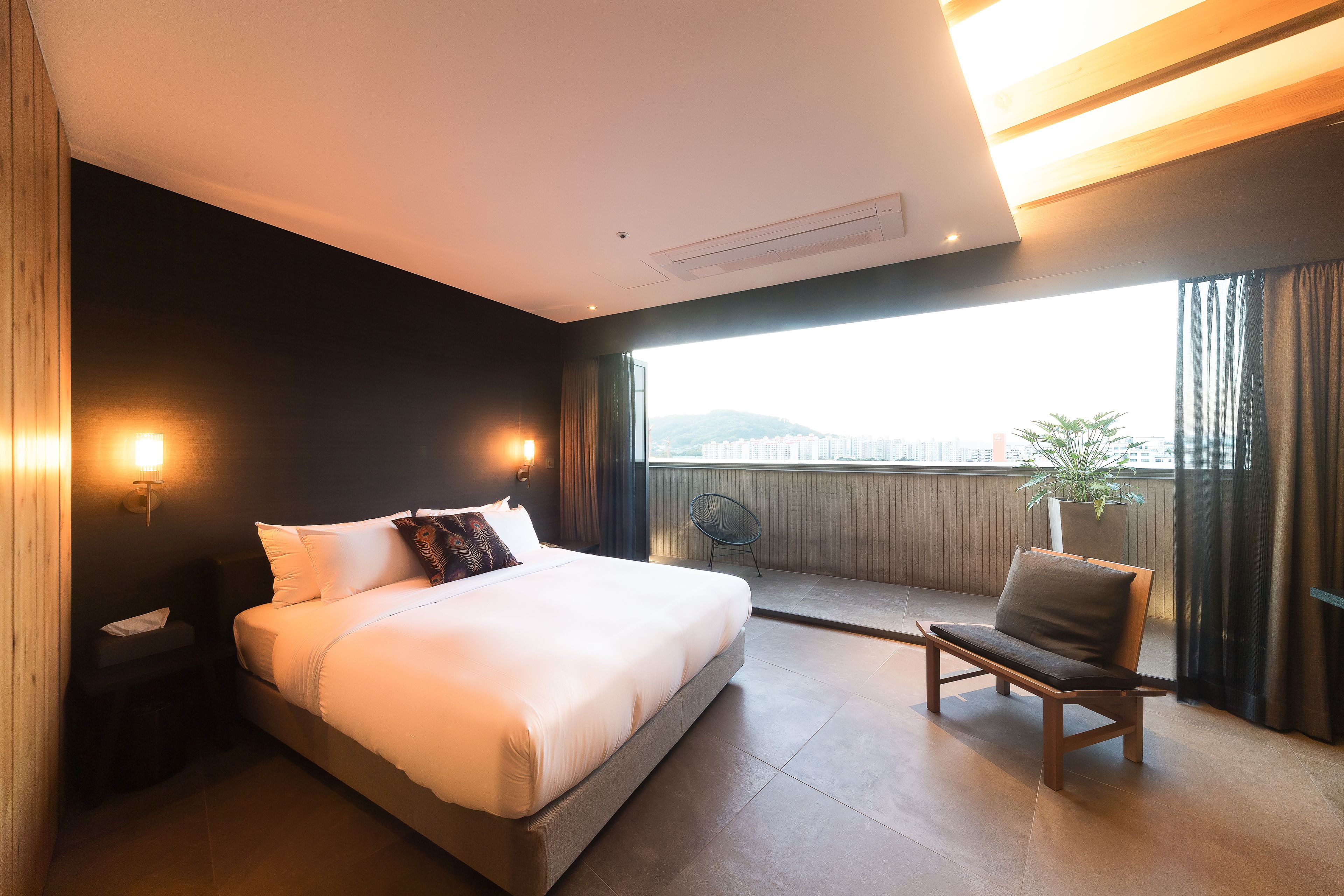 Een moderne hotelkamer met een groot bed, een stoel, een nachtkastje en uitzicht op de stad door een groot raam.