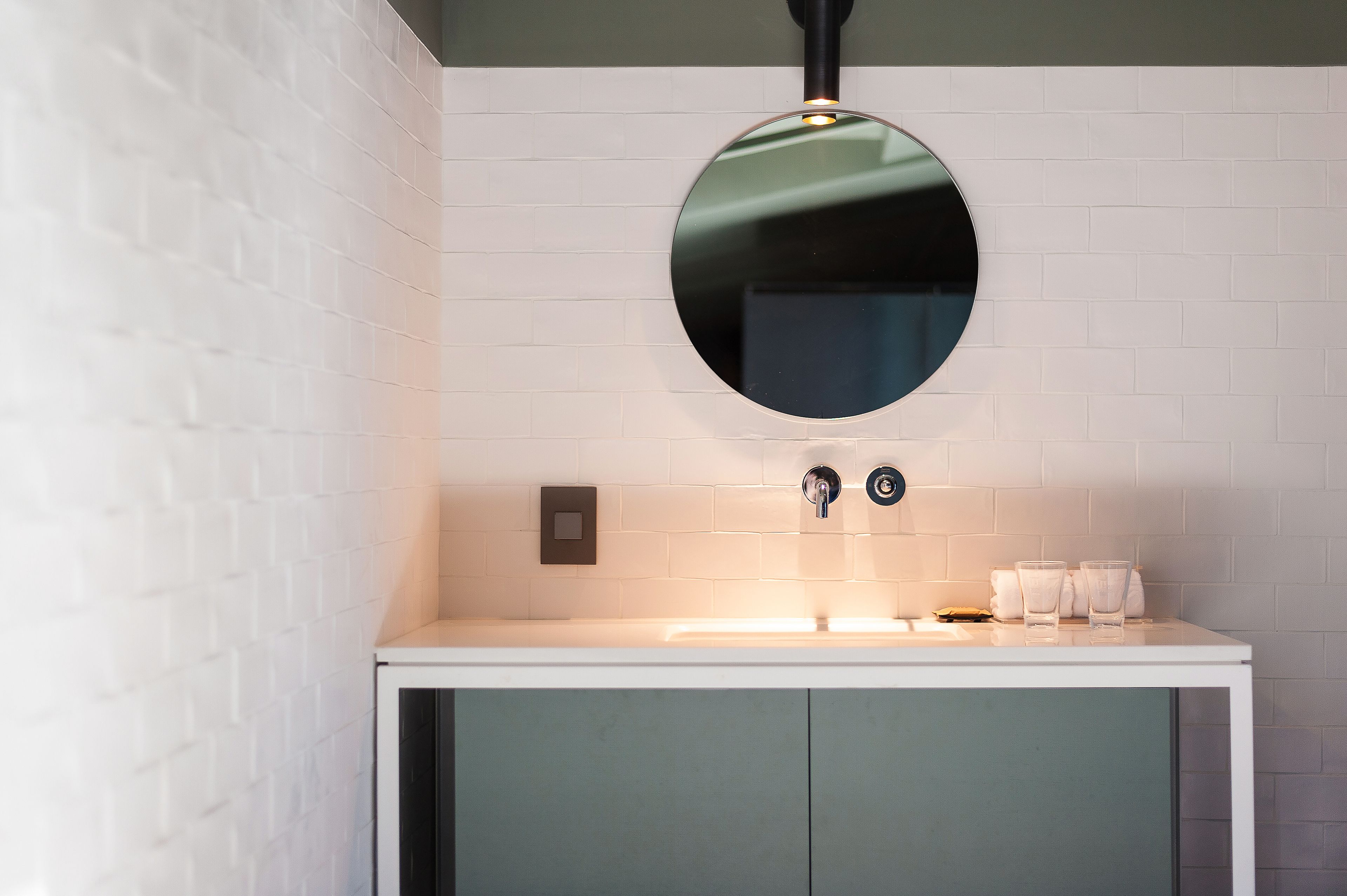 Een moderne badkamer met een betegelde muur, een ronde spiegel, een wit wastafelmeubel en een groene kast.