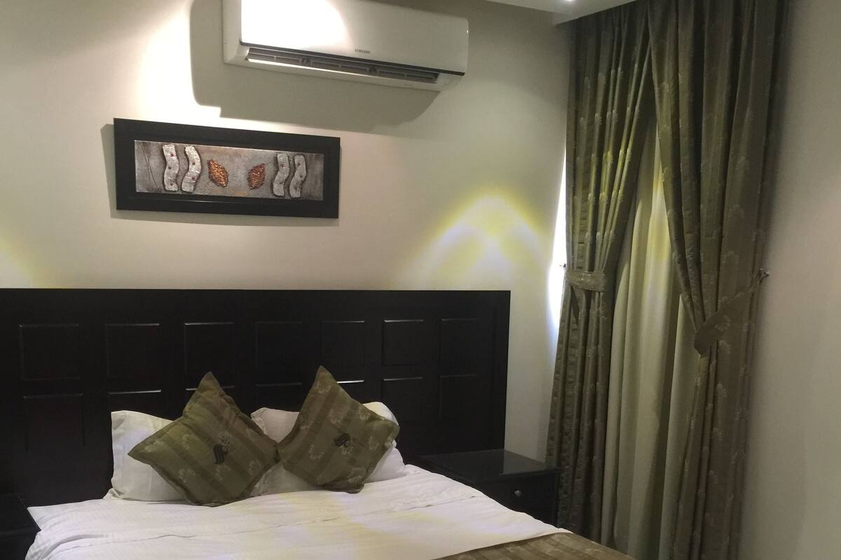 Habitació d'hotel amb un llit gran, un panell de capçalera fosc, un aparell d'aire condicionat, una obra d'art emmarcada i una finestra amb cortines.