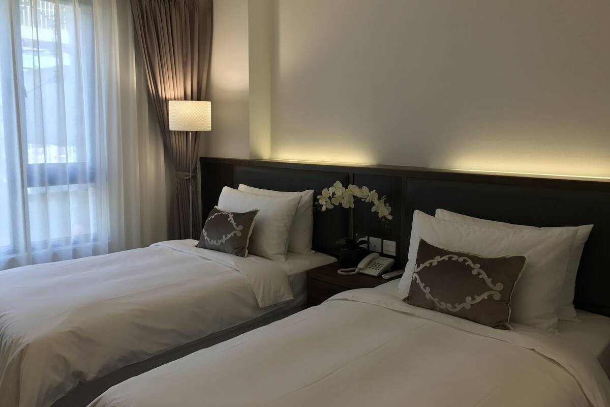 A hotel room with two beds, a nightstand with a phone, and a lamp.