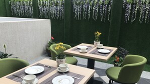 Ein Essbereich mit zwei Tischen, jeder Tisch gedeckt mit Tellern, Tassen und Besteck. Grüne Stühle stehen um die Tische herum, und auf einem Tisch steht ein Topf mit gelben Blüten.