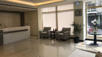A hotel lobby with a reception desk, two patterned armchairs, a potted plant, and a large window.