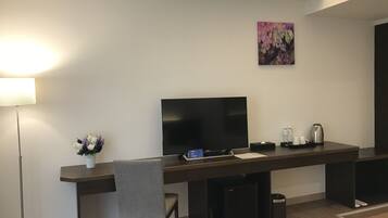 A modern hotel room with a flat-screen TV, a desk with a chair, a small trash bin, and a coffee maker.