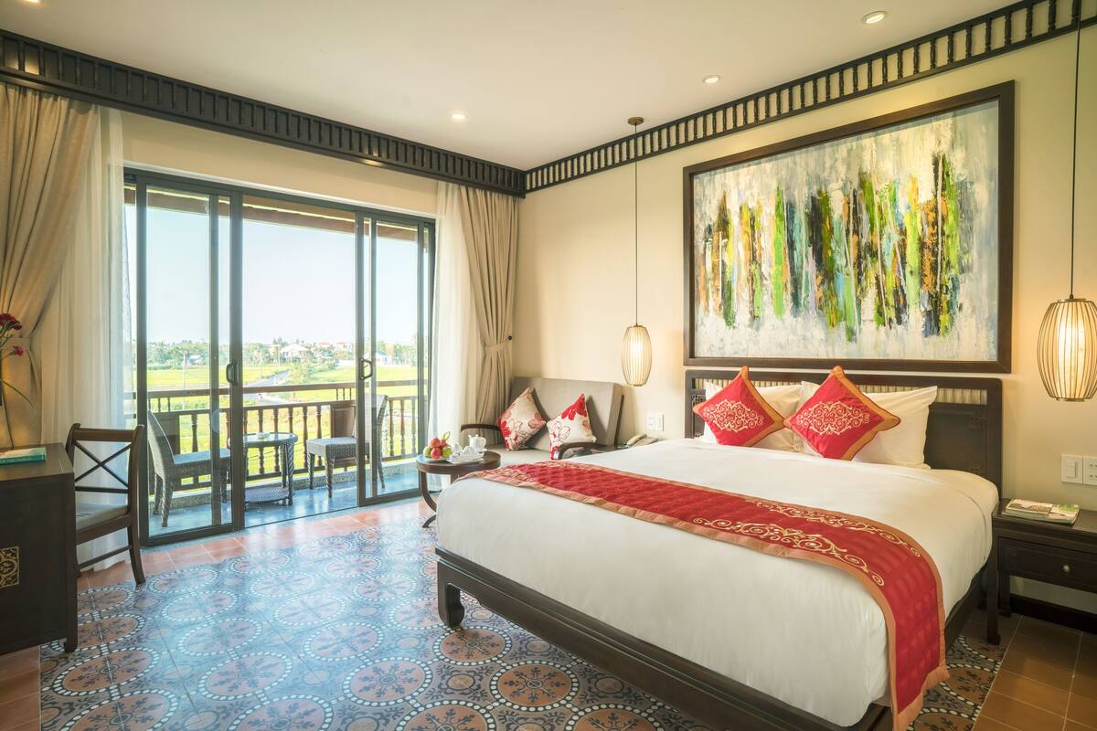 A hotel room with a large bed, a balcony with a view, a painting on the wall, and a desk with a chair.