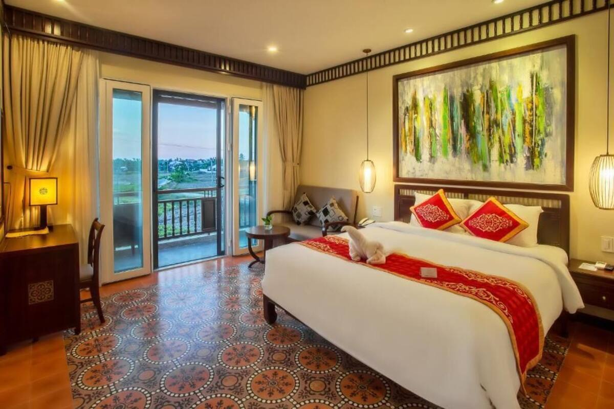 A hotel room with a large bed, a painting on the wall, a balcony with curtains, and a patterned floor.