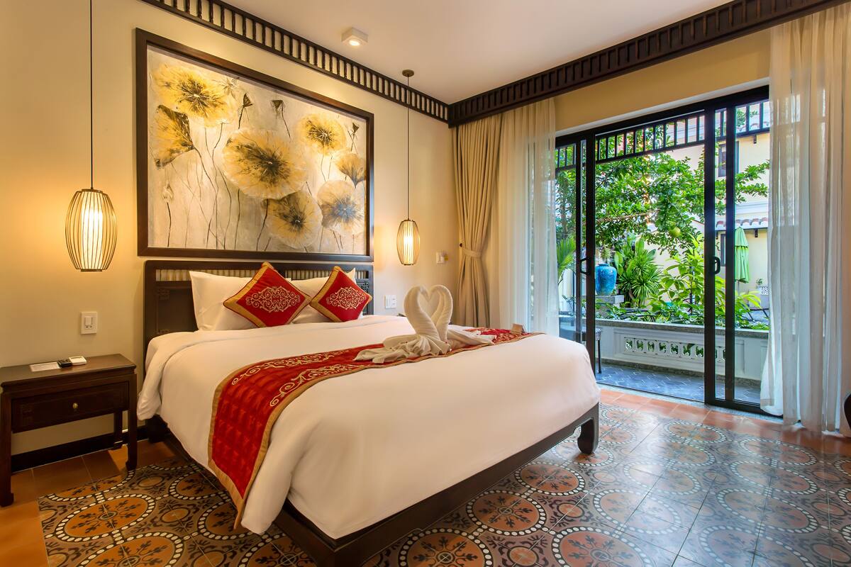 A hotel room with a large bed, a painting on the wall, and a view of greenery outside.
