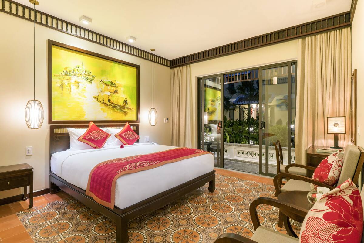 A hotel room with a large bed, a view of the street through a glass door, and a dining area with a table and chairs.