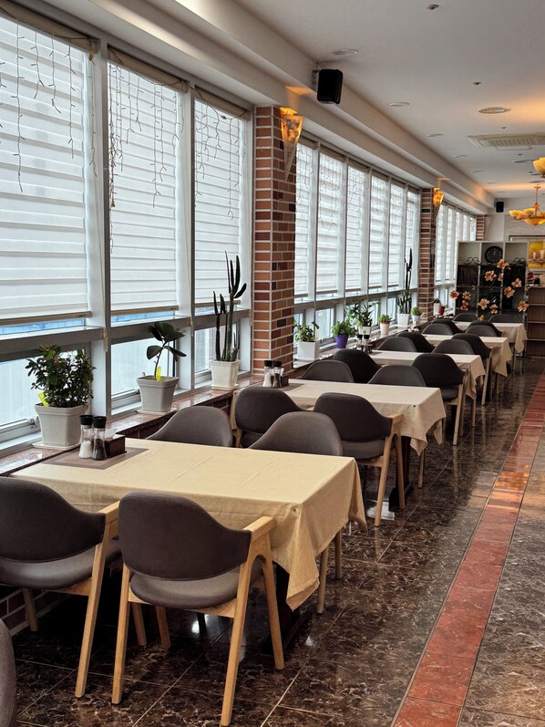 A restaurant with multiple tables and chairs, large windows with blinds, and potted plants.