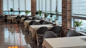 Un comedor amplio con varias mesas y sillas, ventanas grandes con persianas y plantas en macetas.