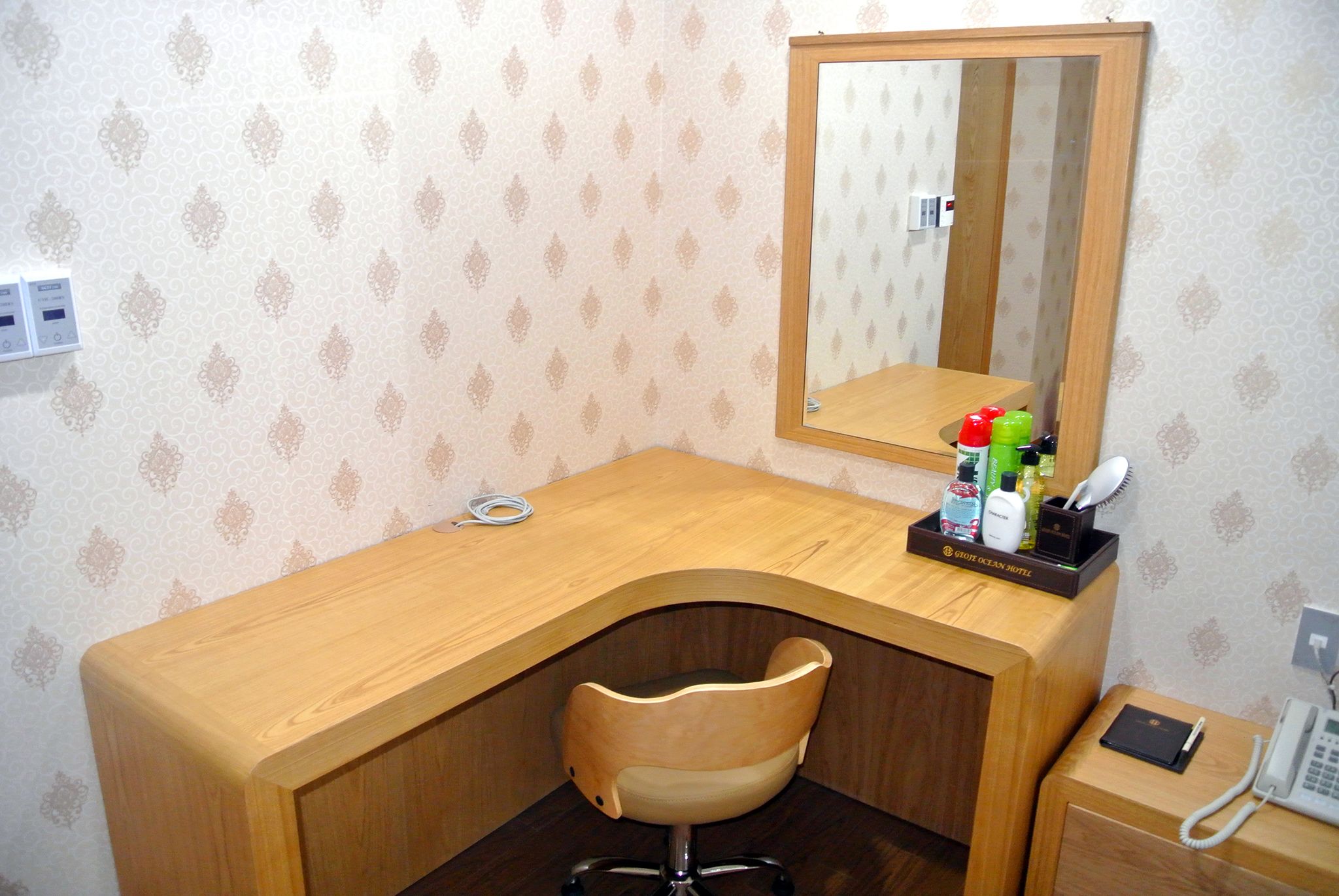 Habitación de hotel con un escritorio de madera, una silla, un espejo, un teléfono y artículos de tocador sobre el escritorio.