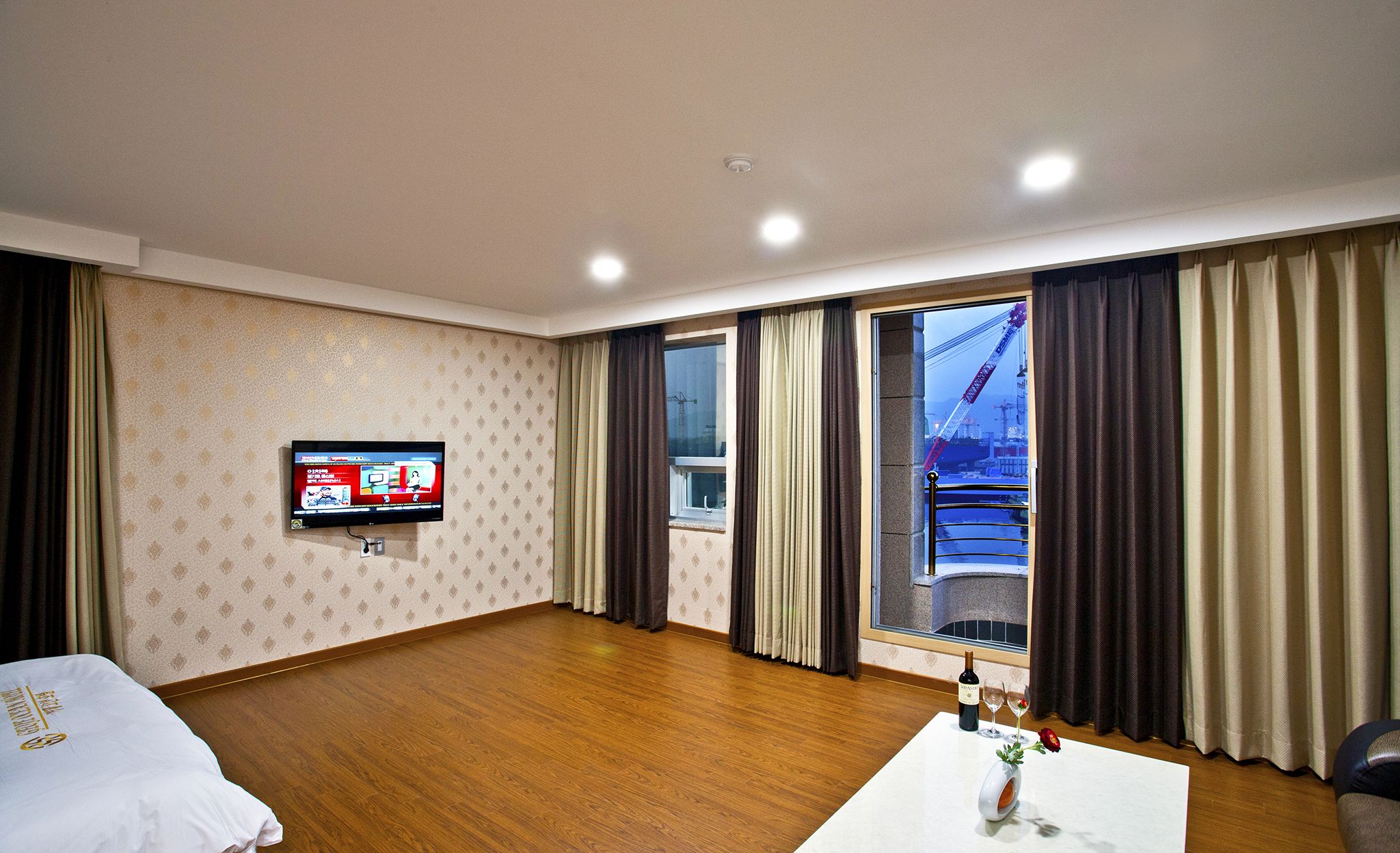 Habitación de hotel con televisión, suelo de madera, cama, ventana con cortinas y una mesa con una botella y vasos.