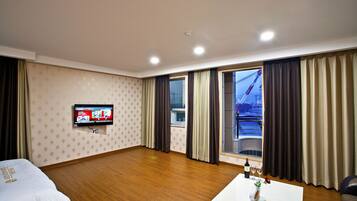 Habitación de hotel con televisión, suelo de madera, cama, ventana con cortinas y una mesa con una botella y vasos.