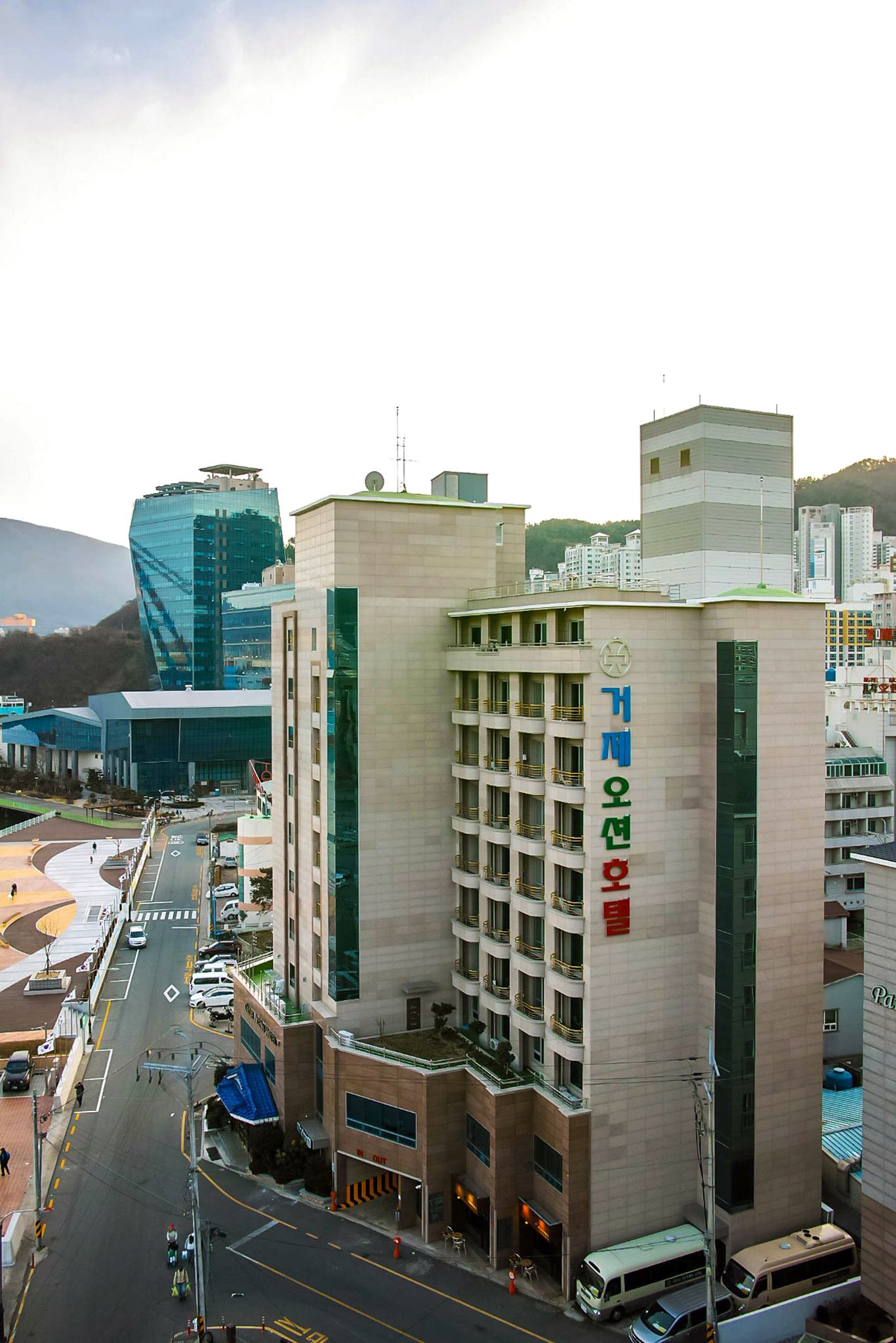 A multi-story hotel with a prominent sign, a parking area, and a modern building in the background.
