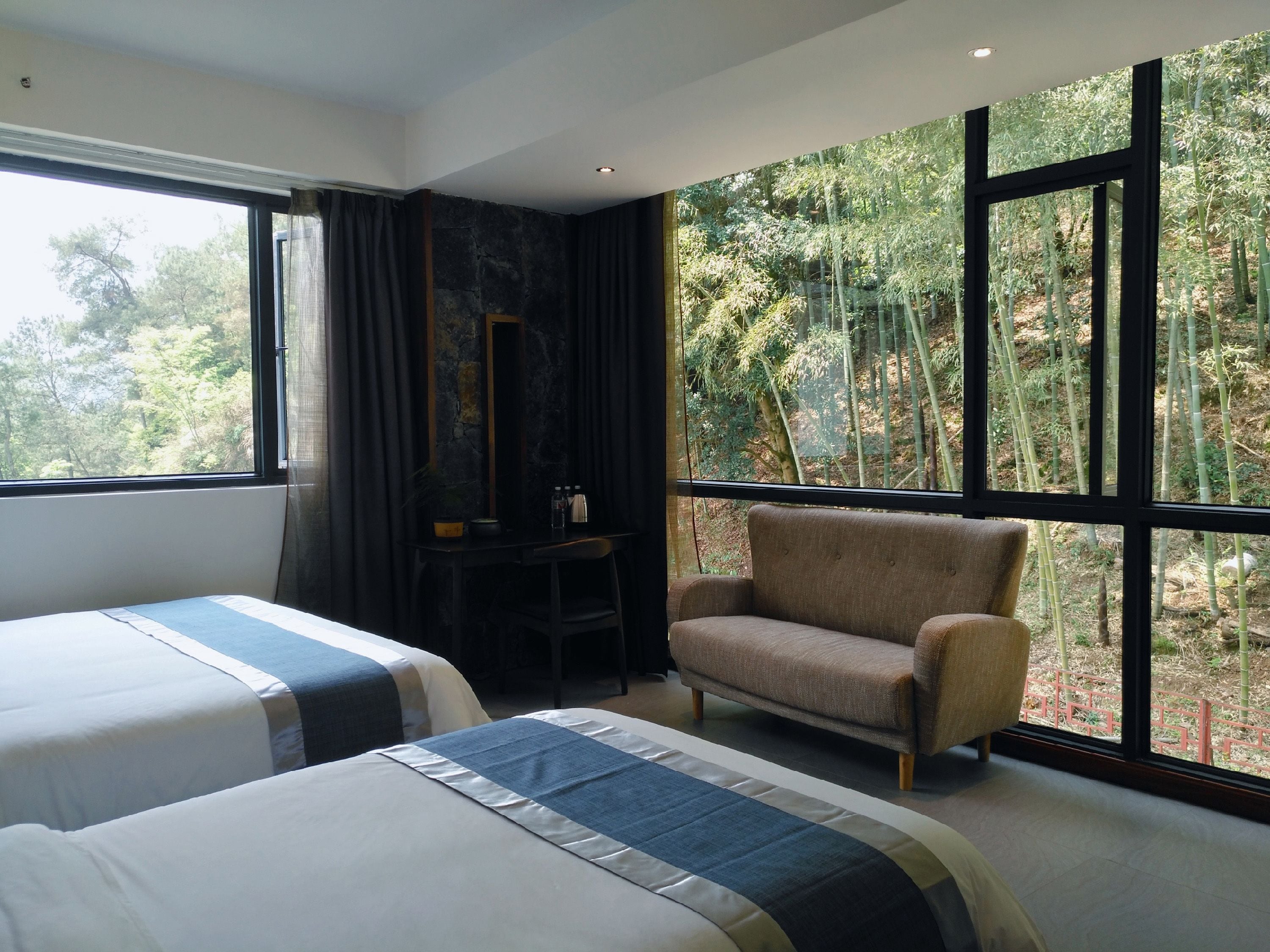 A hotel room with two beds, a chair, a desk, and a large window with a forest view.