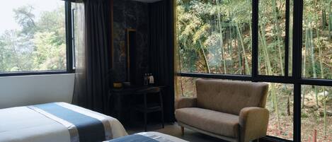 A hotel room with two beds, a chair, a desk, and a large window with a forest view.