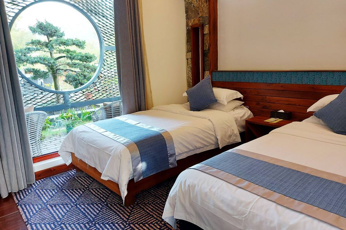 A hotel room with two beds, a view of a tree through a circular window, and a patterned rug.