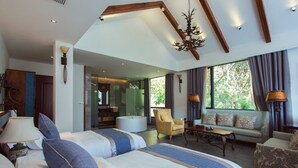 A spacious bedroom with a large bed, a sitting area with a sofa and armchairs, a chandelier, and a view of greenery outside.