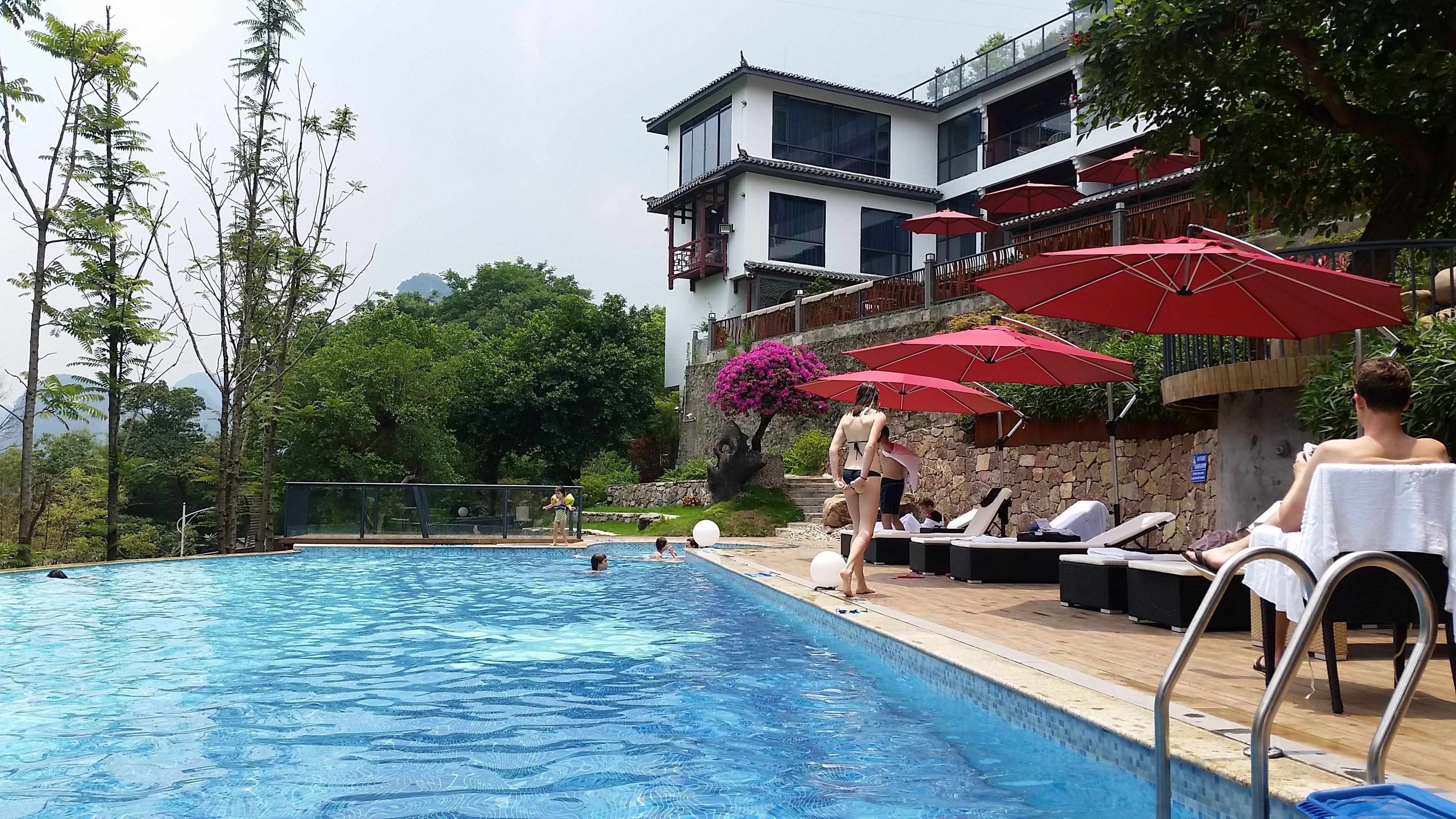 Une piscine entourée d’arbres et d’un bâtiment doté de balcons, avec des chaises longues et des parasols à proximité.