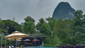 A poolside area with lounge chairs and umbrellas, surrounded by lush greenery and a mountain backdrop. - Li River Resort (Guilin)