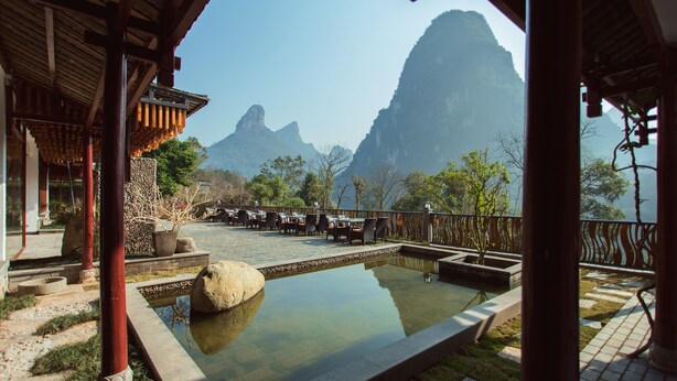 A traditional wooden building with a pool, outdoor seating, and a view of mountains. - Li River Resort (Guilin)