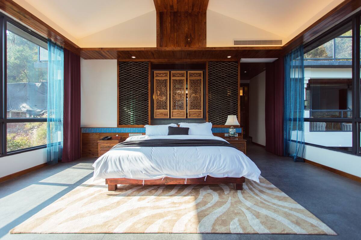 A bedroom with a large bed, wooden headboard, and a view of the outdoors through large windows.