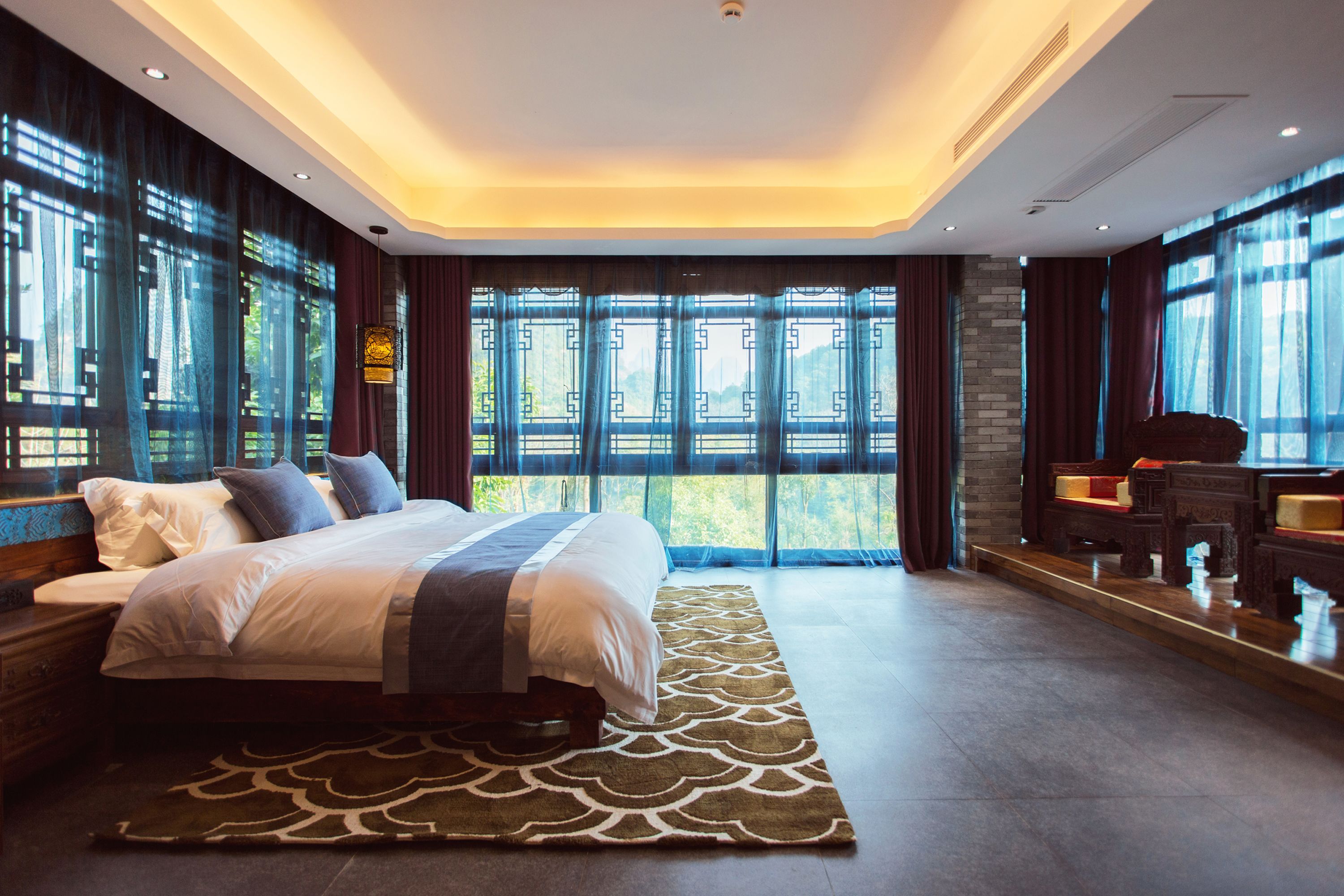 A modern hotel room with a large bed, a seating area with a table, and a view of the outdoors through large windows.