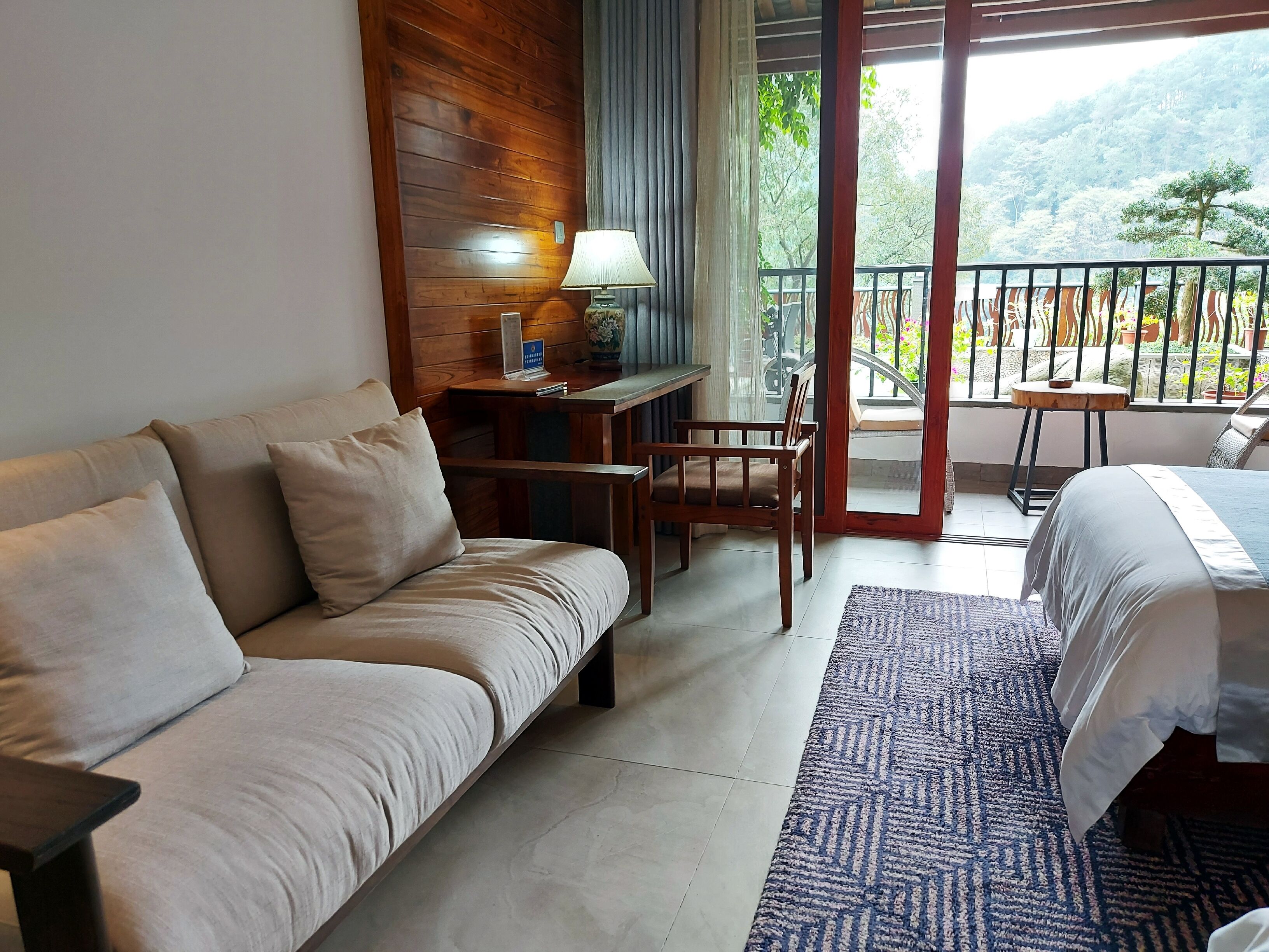 A hotel room with a sofa, a bed, a small table with a lamp, and a view of a balcony with outdoor furniture.