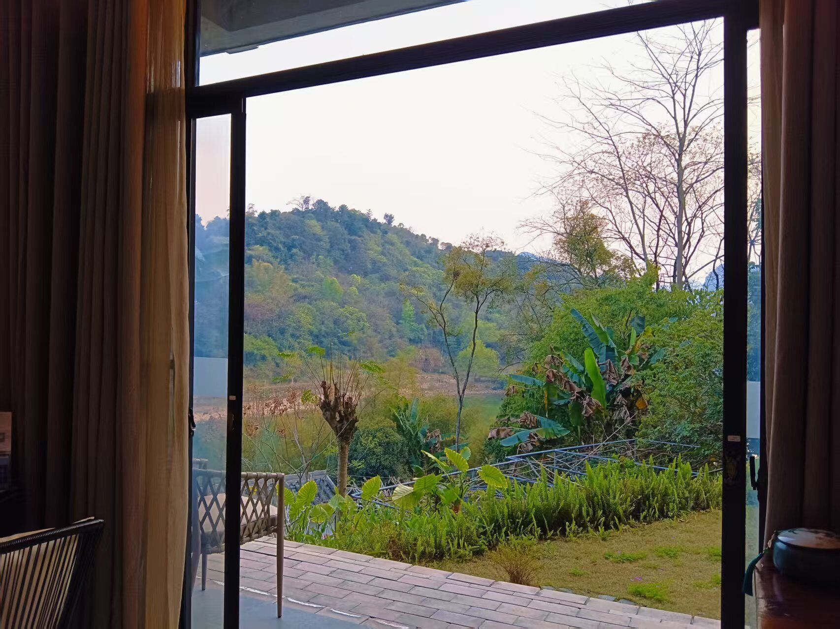A room with a large window overlooking a lush garden and a hilly landscape.
