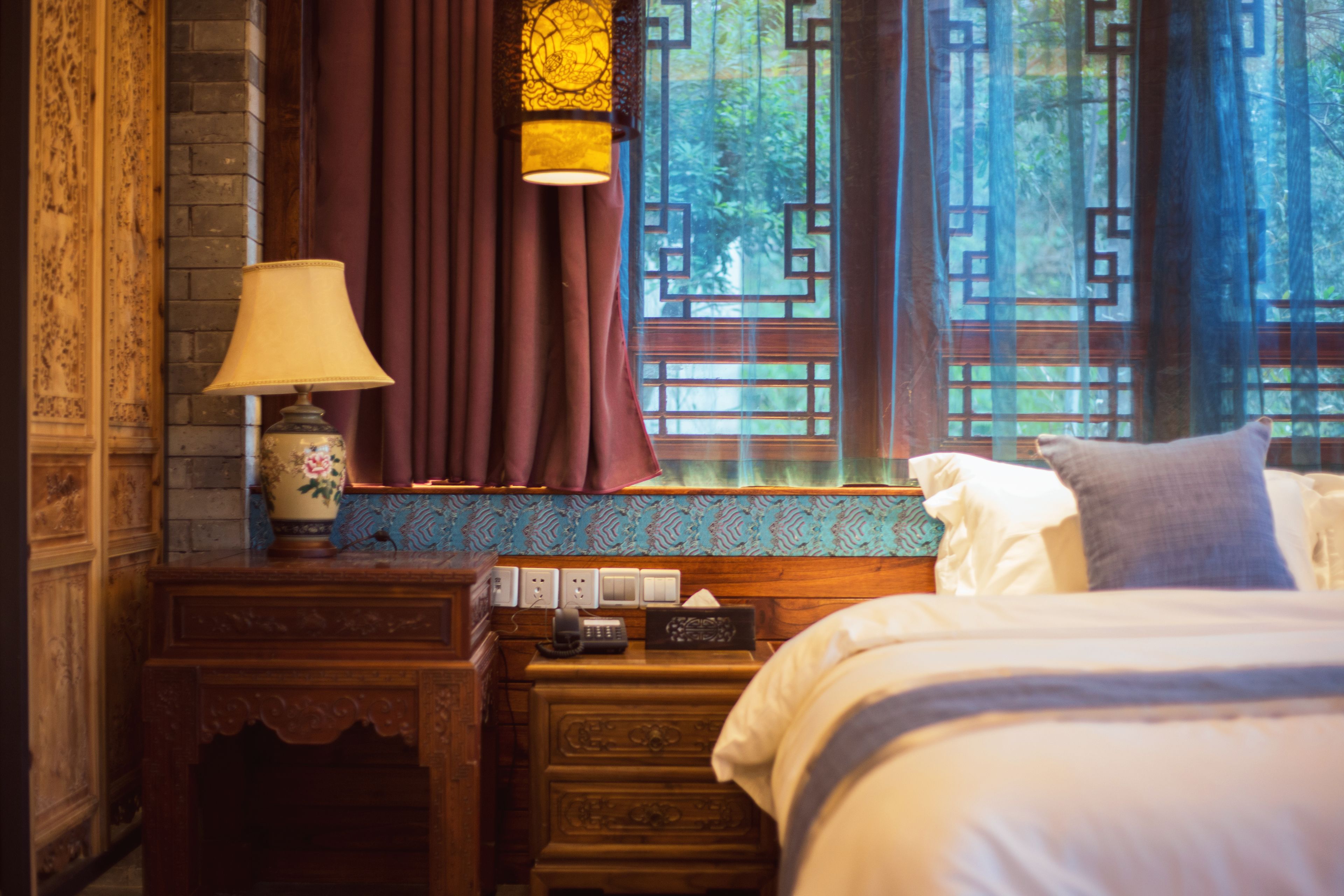 A traditional room with a bed, a nightstand, a lamp, and a window with decorative curtains.
