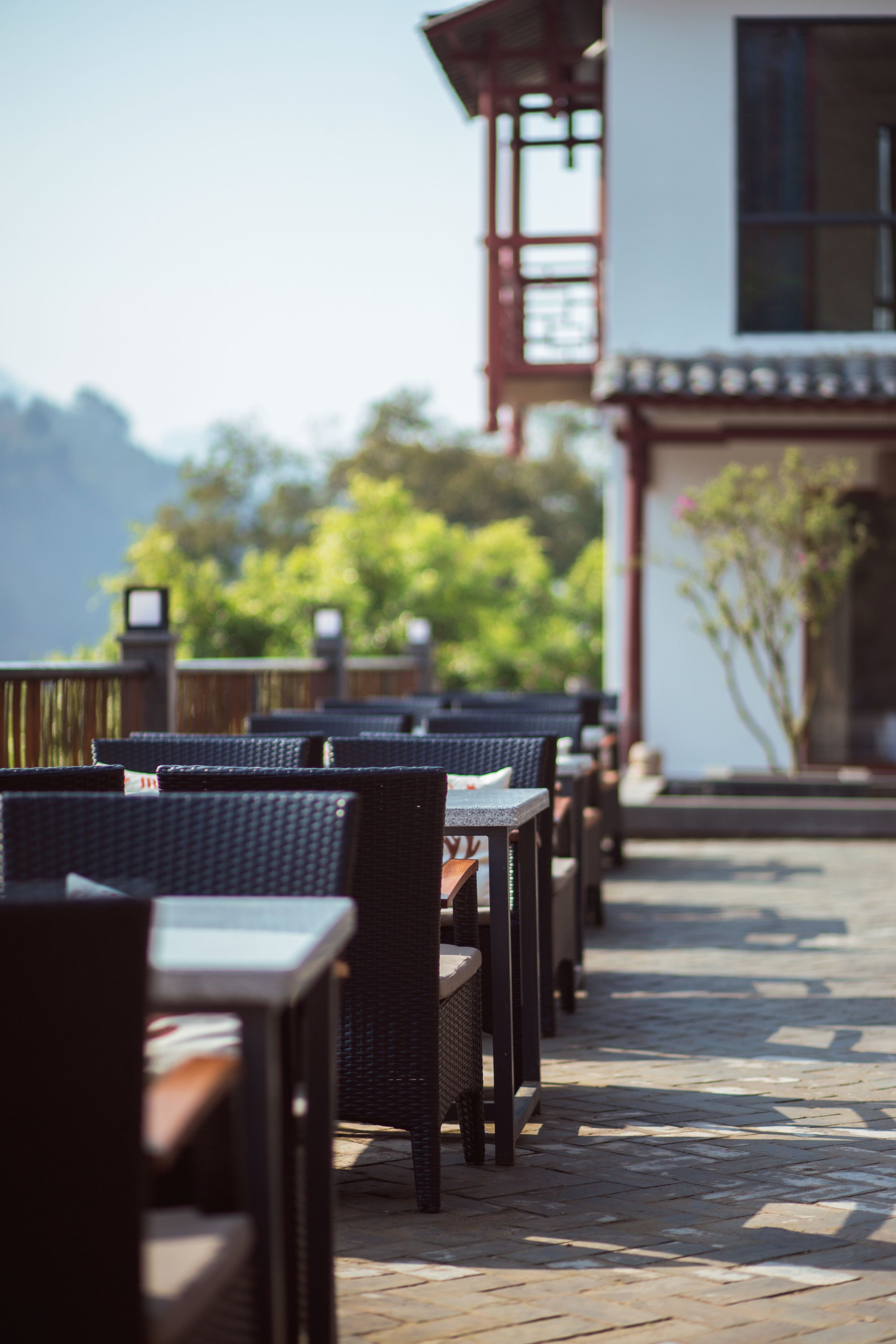 Des tables à manger en plein air, accompagnées de chaises en osier, une terrasse en bois et, au fond, un bâtiment présentant un style architectural traditionnel.