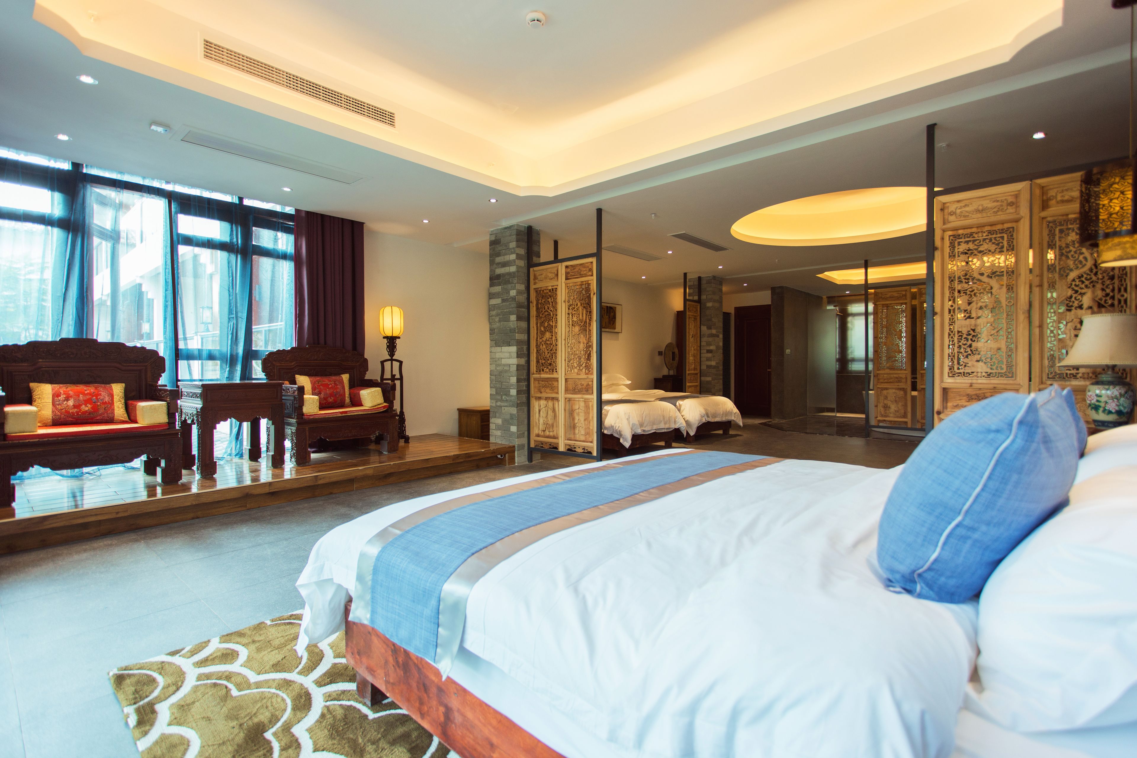 A spacious hotel room with a large bed, a sitting area with a sofa and chairs, a large mirror, and a view of the outside through large windows.