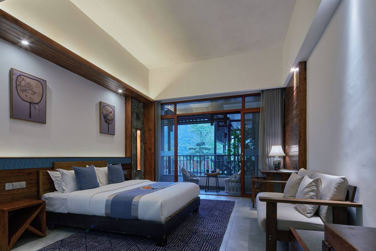 A modern hotel room with a large bed, a seating area, and a balcony with a view of trees.