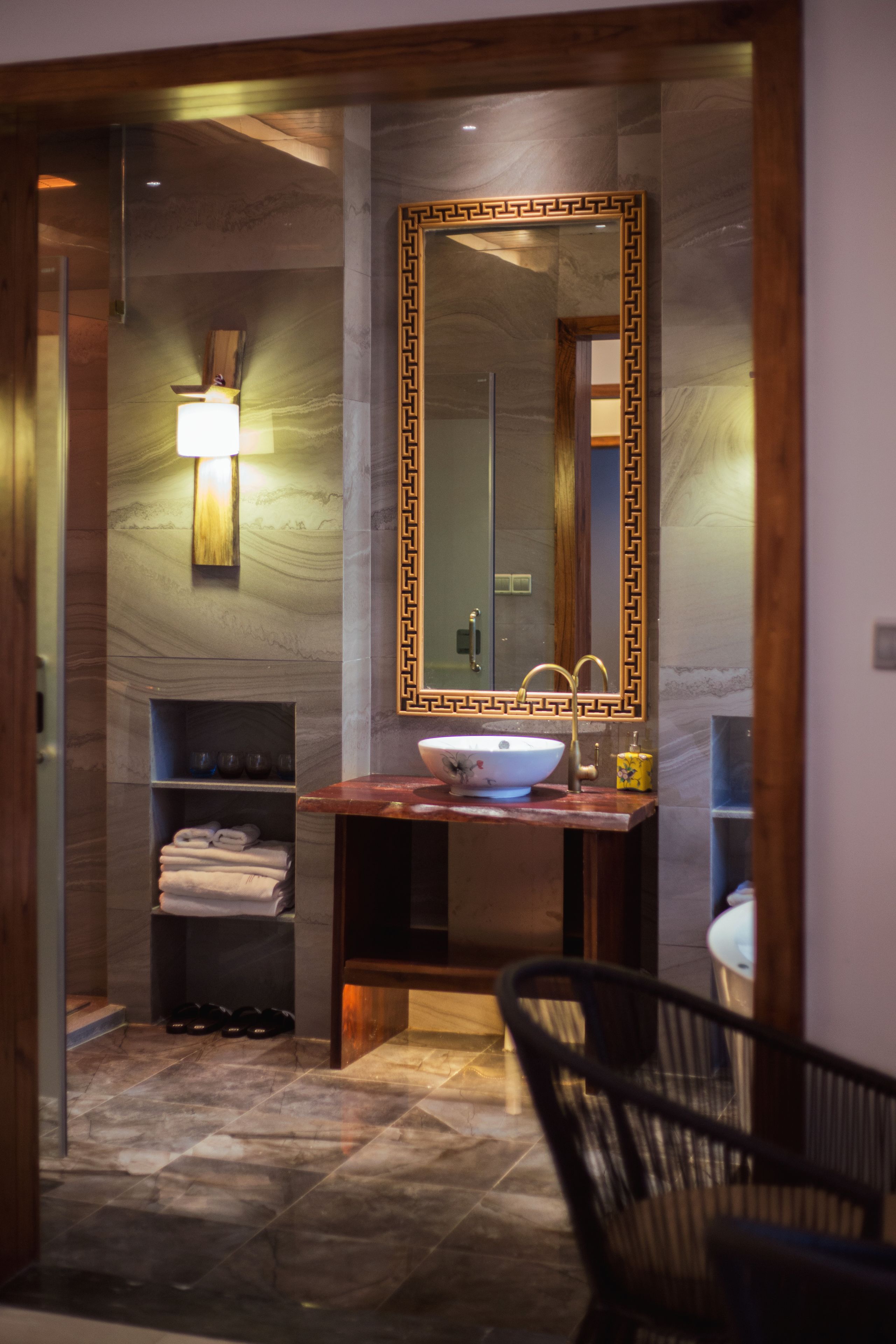 Un baño con pared de mármol, un espejo grande con marco decorativo, un lavabo con recipiente y un perchero con toallas dobladas.
