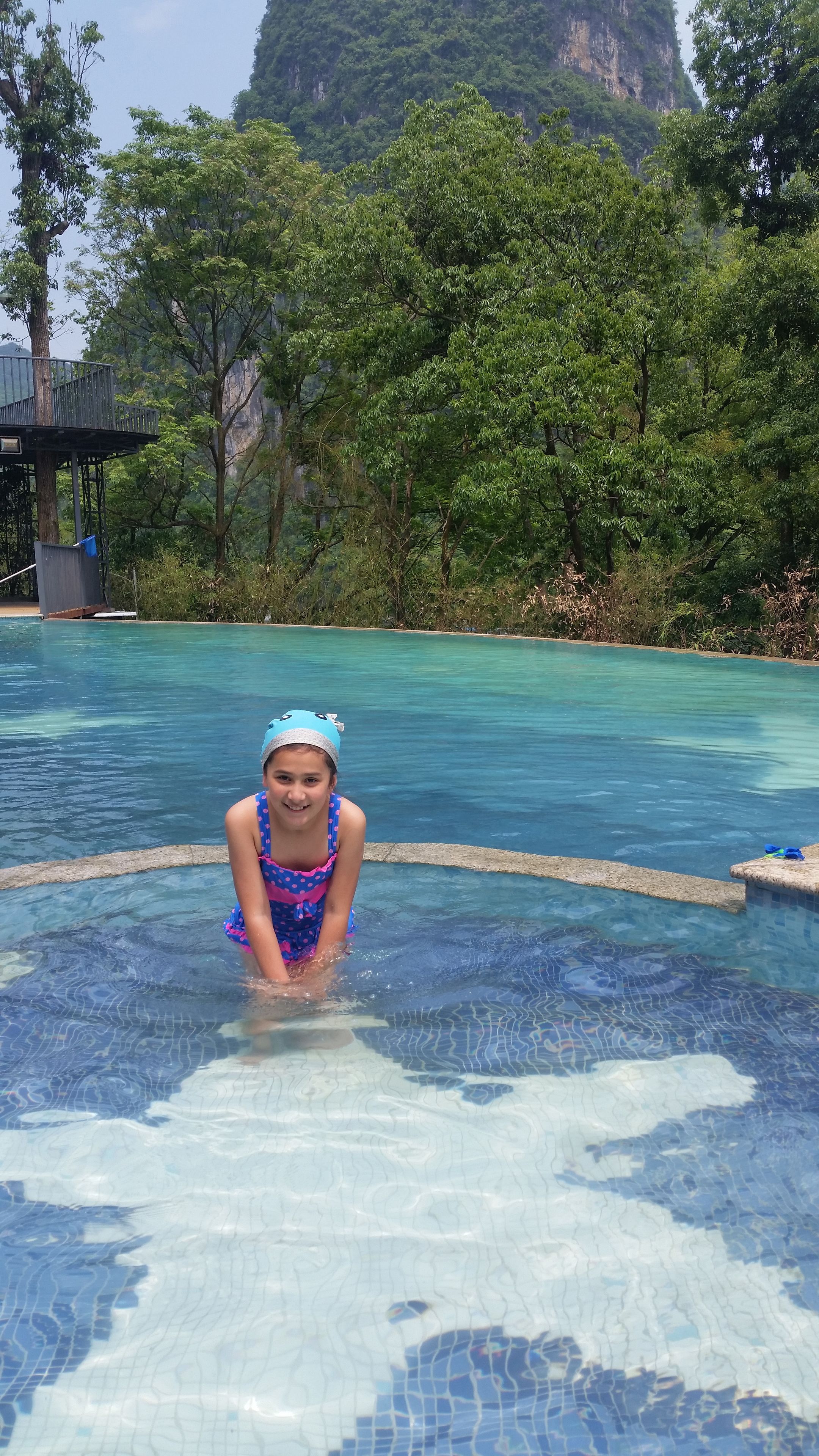 A person in a swimming pool with clear blue water, surrounded by lush greenery and a mountainous backdrop.