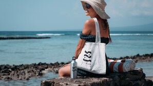 A person sitting on a rock by the sea, holding a water bottle and a white tote bag with "UTARA VILLAS" printed on it. - Utara Villas (Gili Trawangan)